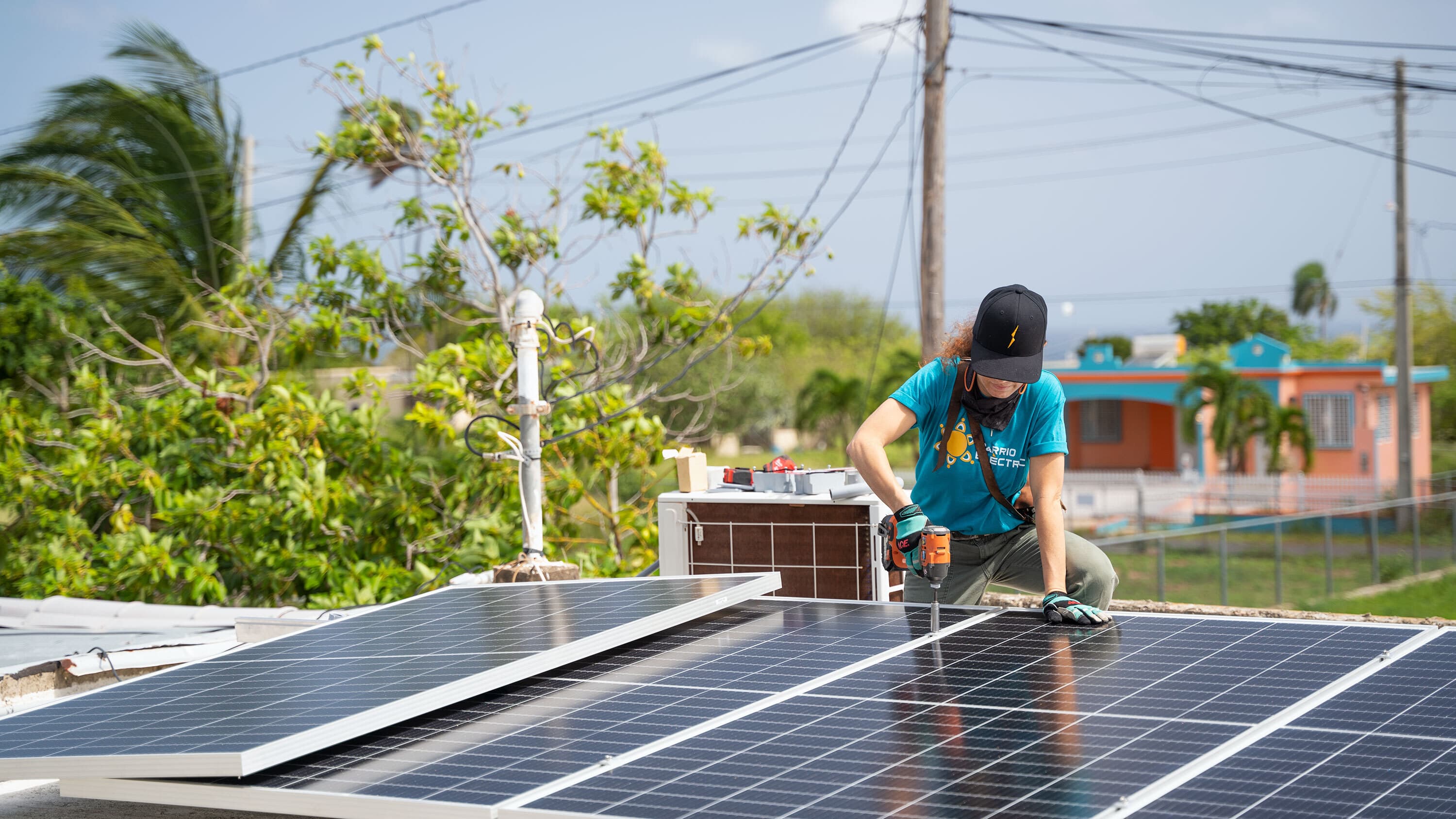 A person wearing a hat and gloves is installing solar panels on a rooftop in a residential area, using a power drill. Trees, power lines, and colorful houses are visible in the background.