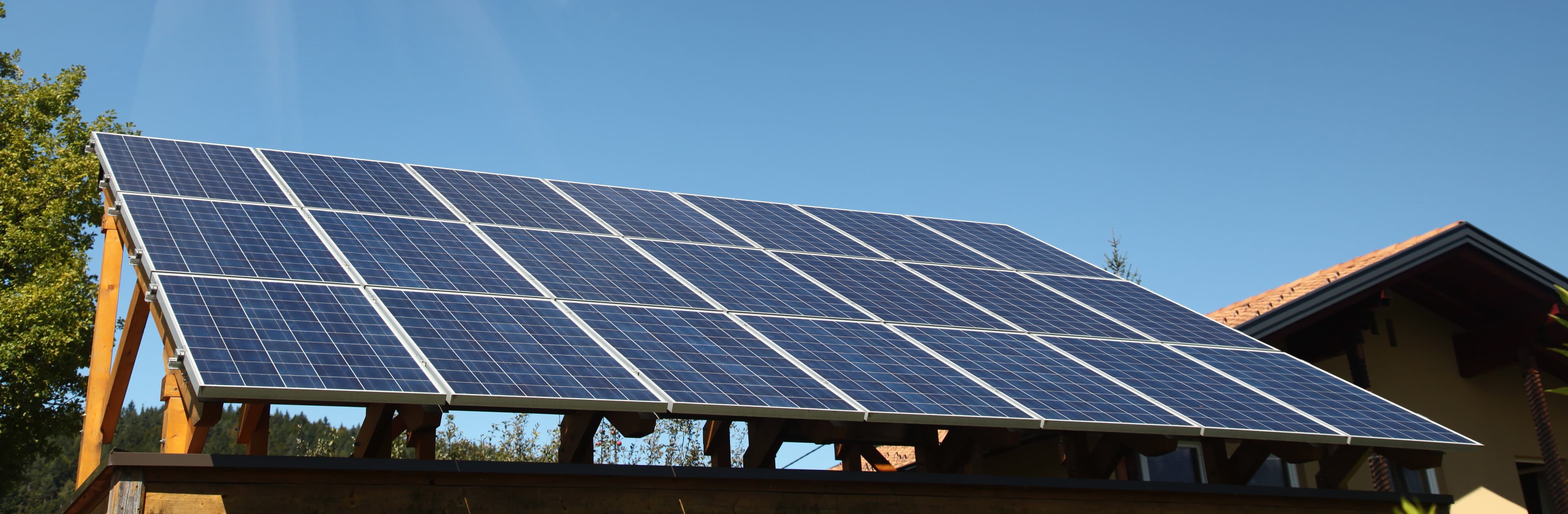 A rooftop solar panel array is mounted on a wooden structure under a clear blue sky, with part of a house visible in the background.