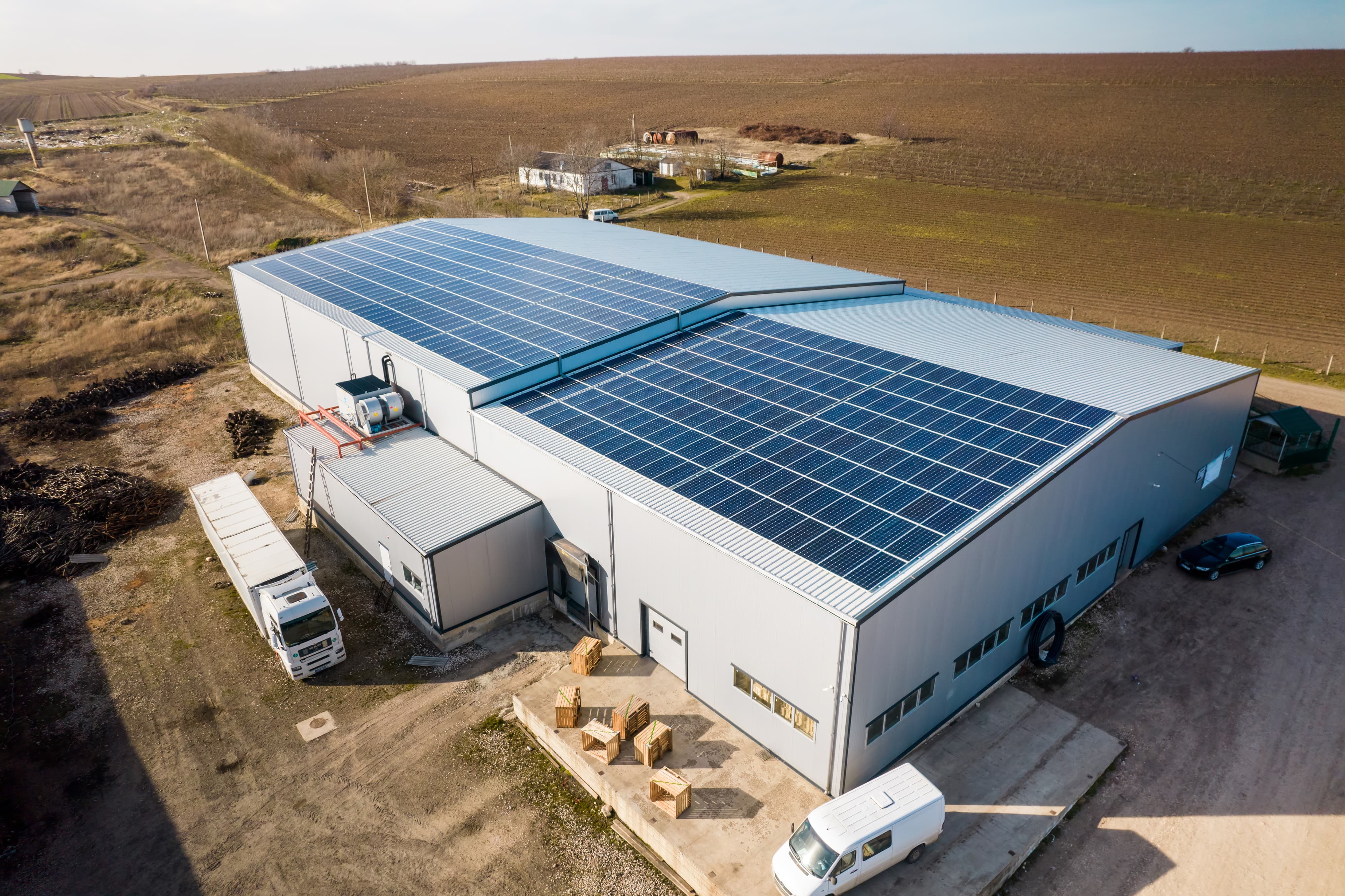 An industrial warehouse in a rural area is equipped with a large array of solar panels on its roof. Surrounding the building are trucks, a van, and open fields.