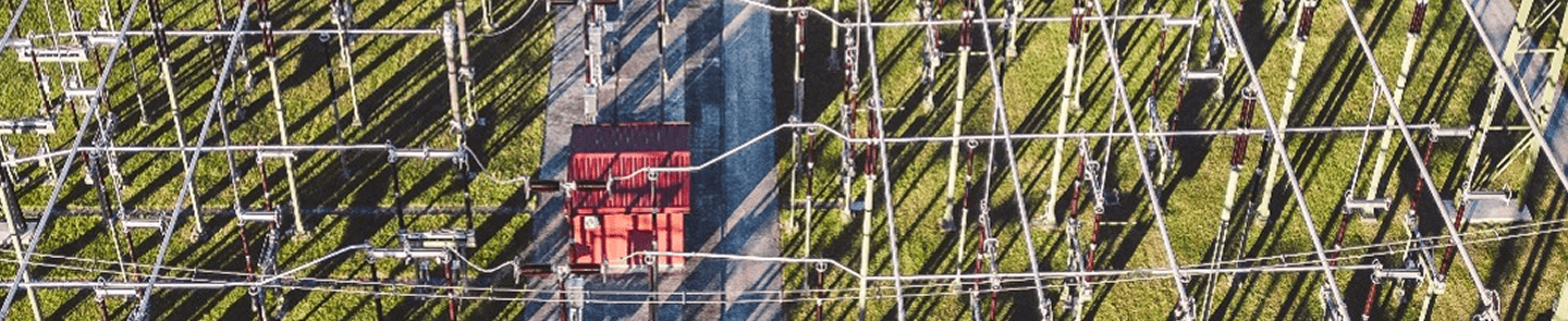 Aerial view of an electrical power grid substation with a red-roofed control building, metal framework, and high-voltage equipment arranged in a grid pattern on green grass.